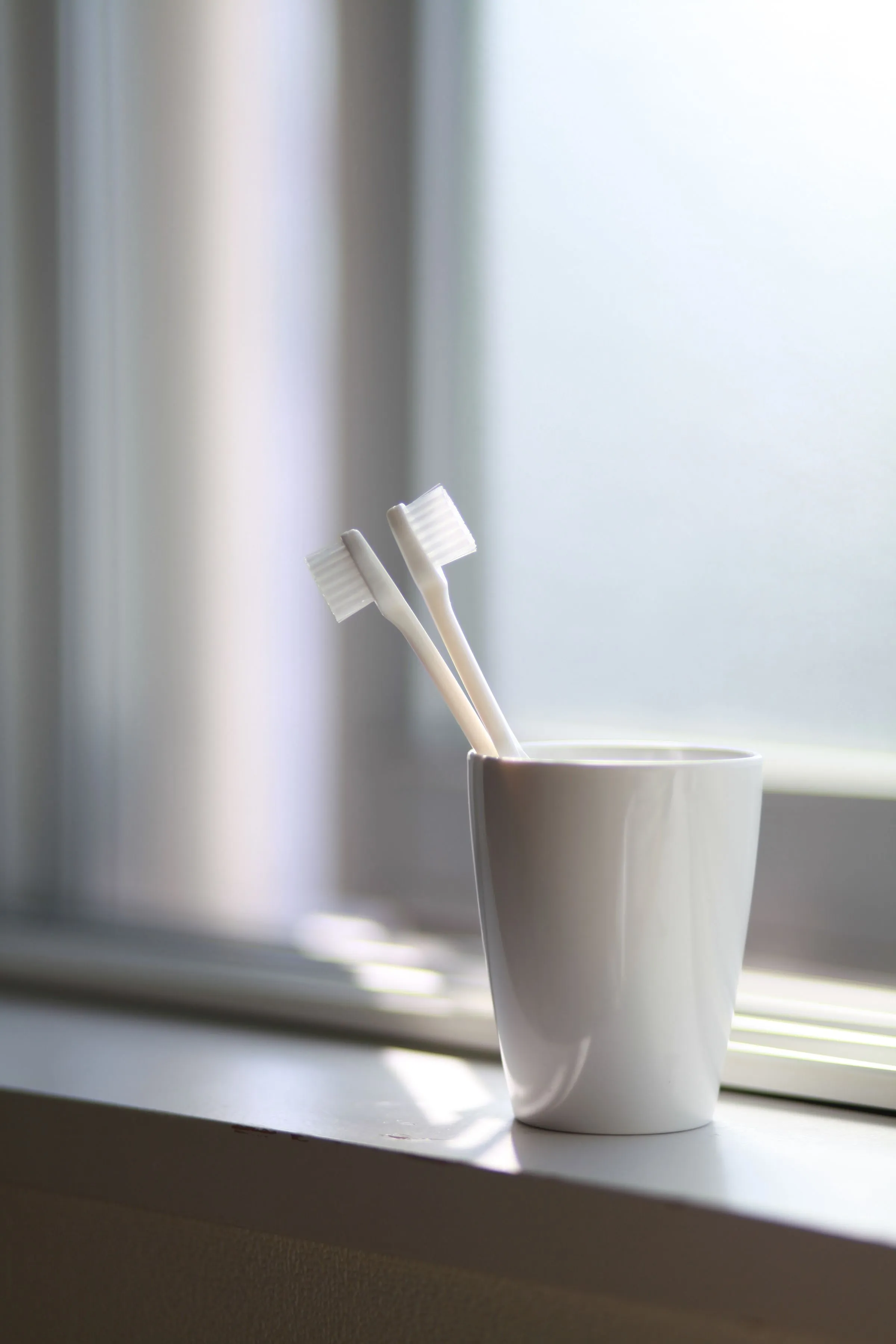 Two white toothbrushes in a ceramic cup on a sunny windowsill